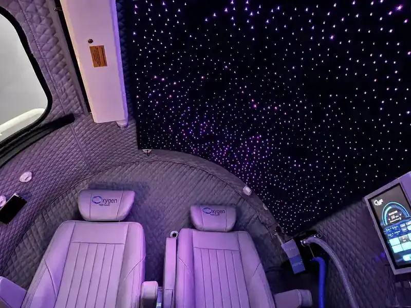 Interior view of the M5500 chamber's fiber-optic starlight ceiling glowing with purple star points above two lavender quilted-leather seats with embroidered Oxygen Health Systems headrests and the touchscreen control panel